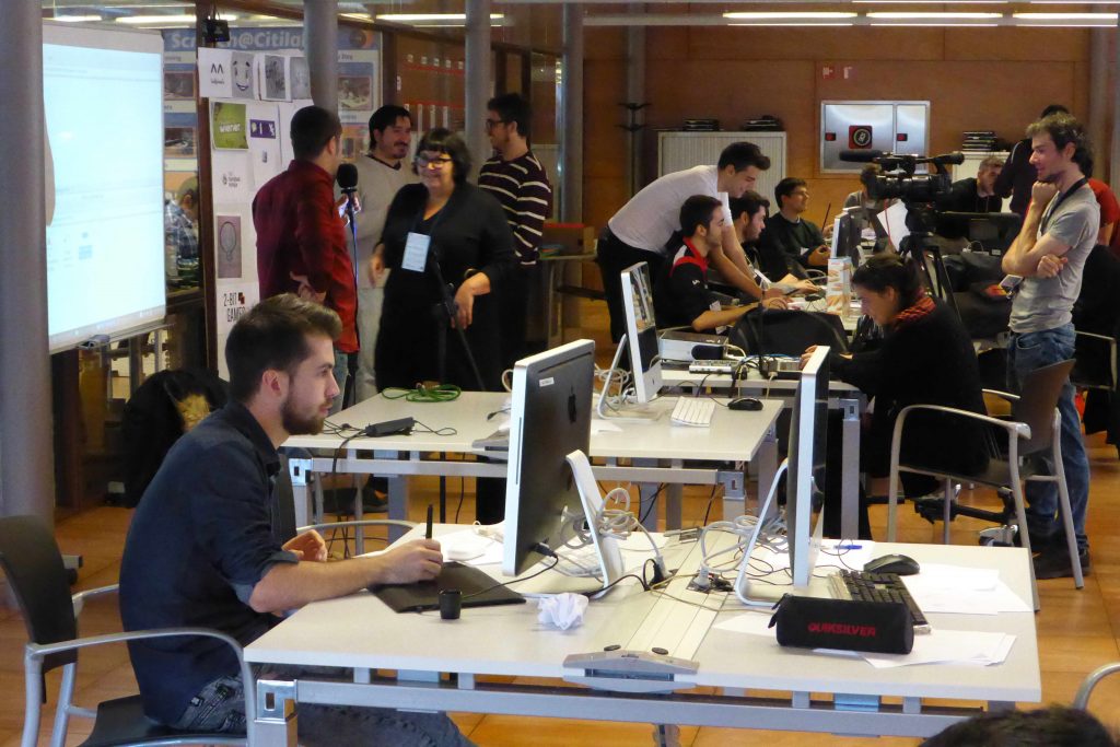 Un grup de persones treballant amb ordinadors en una oficina moderna, algunes col·laborant en jocs educatius, altres concentrades en pantalles, mentre un equip de càmeres enregistra l'atmosfera concorreguda i col·laborativa.