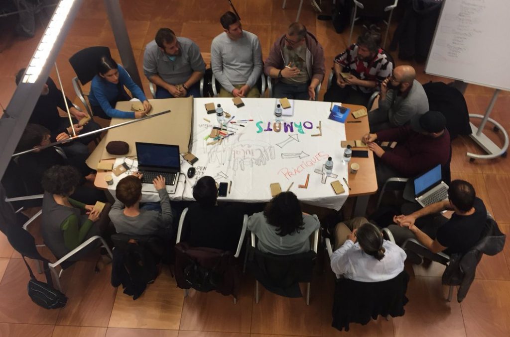 Un grup de persones seuen al voltant d'una gran taula coberta de papers, ordinadors portàtils i tasses de cafè, immerses en una conversa durant la Setmana de Cocreació de Cornellà. Les lletres de colors a la taula escriuen "TOMMOS" i les fletxes apunten a elements etiquetats.