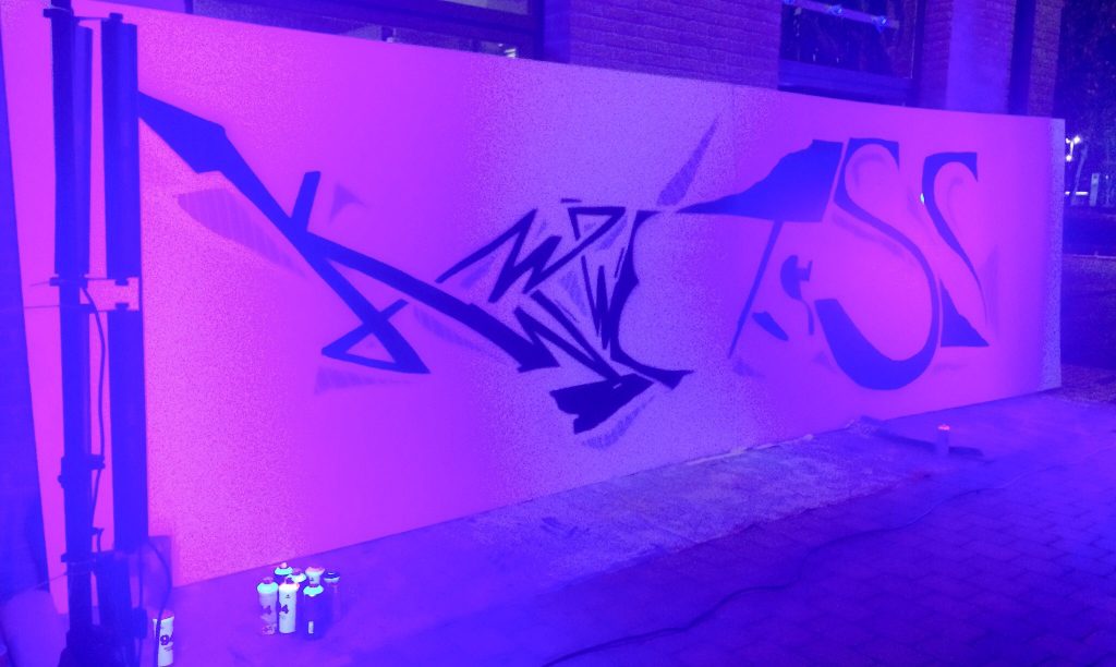 Una paret coberta de grafits negres abstractes, il·luminada per una llum morada brillant, commemora un desè aniversari. Diversos pots de pintura en esprai hi ha a terra, amb el paviment i part d'un edifici visibles al fons.