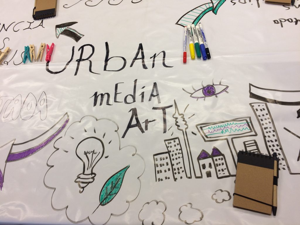 Una taula coberta amb paper blanc mostra dibuixos de colors d'edificis, una bombeta i les paraules URBAN media ART. Marcadors de colors i blocs de notes petits estan escampats per la taula durant la Setmana de Cocreació de Cornellà.