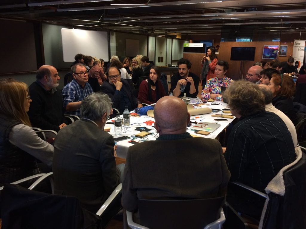 Un grup de persones seuen al voltant d'una taula debatent durant la Setmana de Cocreació de Cornellà, amb papers i materials escampats, en una sala ben il·luminada i plena d'altres participants.