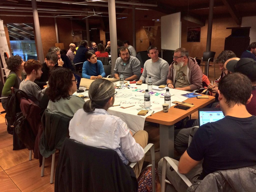Un grup de persones seuen al voltant d'una gran taula en una sala de reunions durant la Setmana de Cocreació de Cornellà, immerses en una discussió. Papers, retoladors i ampolles d'aigua són sobre la taula. Diversos prenen notes mentre una persona treballa amb un ordinador portàtil.