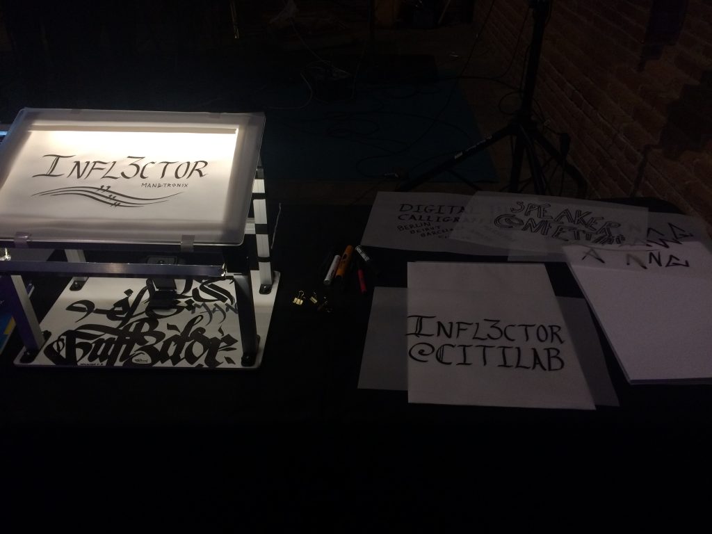Una taula mostra una caixa de llum amb la paraula INFLECTOR en lletres estilitzades, envoltada de papers amb diversos dissenys de cal·ligrafia escrits a mà i alguns retoladors, creant un ambient acollidor que recorda una celebració d'un desè aniversari.