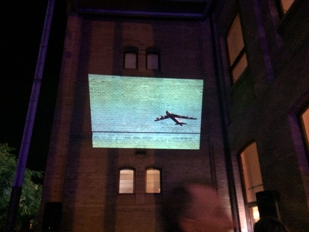 Una projecció d'un avió volant es mostra a la paret de maons d'un edifici a la nit durant el desè aniversari, amb algunes persones i finestres visibles en primer pla.