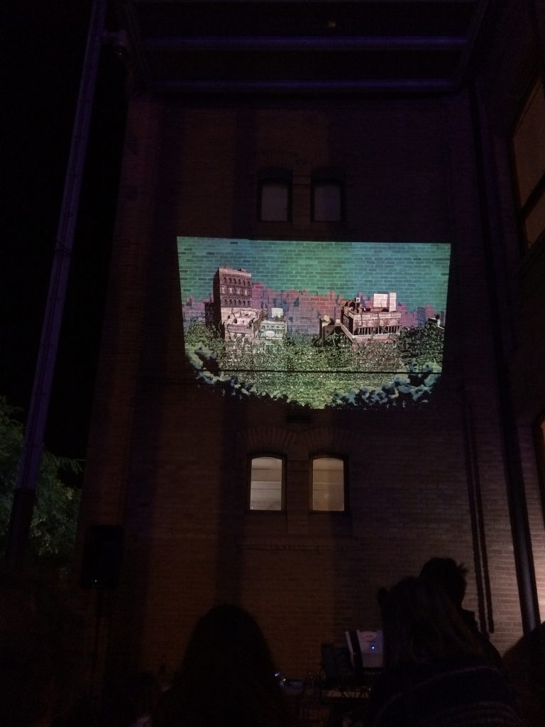 Una projecció nocturna a l'aire lliure per a la Setmana de Cocreació de Cornellà mostra un paisatge urbà ple de color sobre la paret d'un edifici de maó, amb siluetes de gent mirant en primer pla. Sota la vibrant projecció es poden veure dues finestres.
