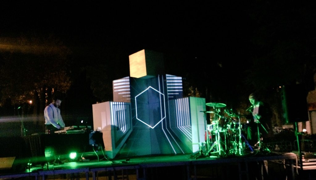 Un escenari nocturn a l'aire lliure presenta una instal·lació de llum geomètrica al centre, que brilla en verd i blanc per al desè aniversari. Un DJ es troba a la consola esquerra, mentre que un bateria actua a la dreta, tots dos envoltats de llum verda.
